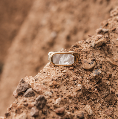 The ring "Shell Signet" from Triada Designs is a golden ring with a mother of pearl shell signet. it lyes on warm earthy texture