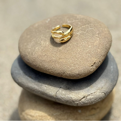 A gold ring from Triada Designs, called "Open Waves" is lying on three round stones