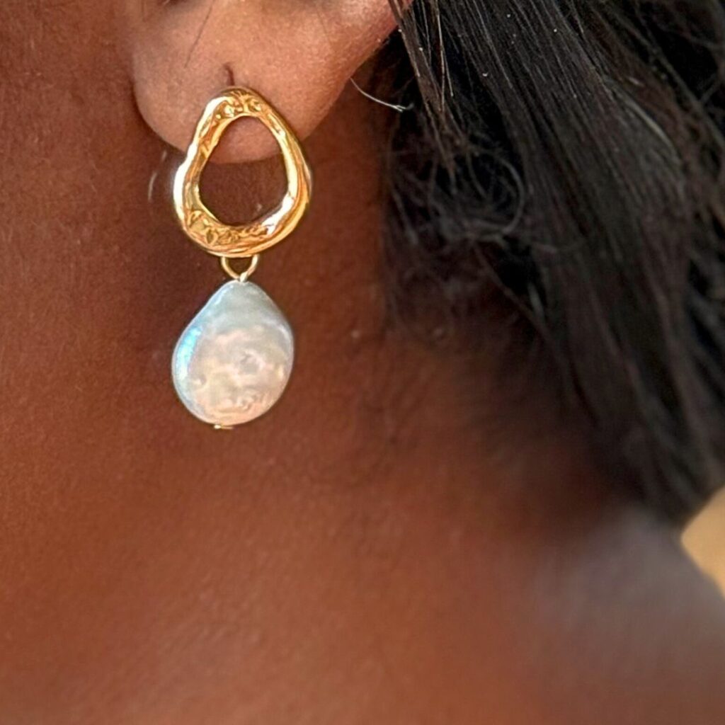 A golden irregular huggie with a fresh water pearl is hanging from the ear of Thekla Matei – An earring from the collection Salt and Spirit, called "Tides Treasure"