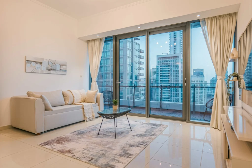 Elegant, modern room with neutral interior design. A couch with a coffetable, a television and a huge arched window – showing the skyscrapers of Dubai.